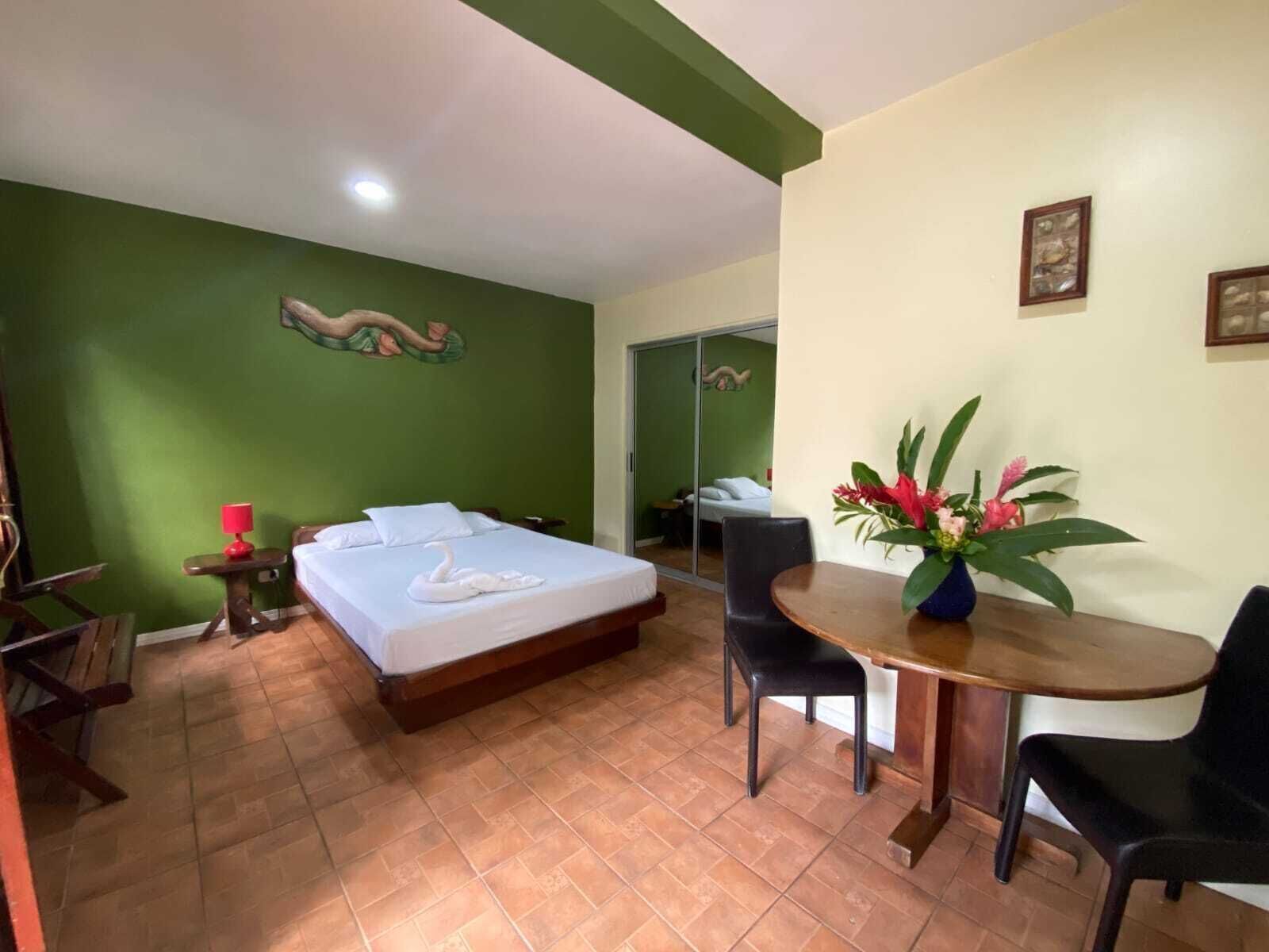A hotel room with a bed, a dining table with chairs, a vase of flowers, and a green wall with a mural.