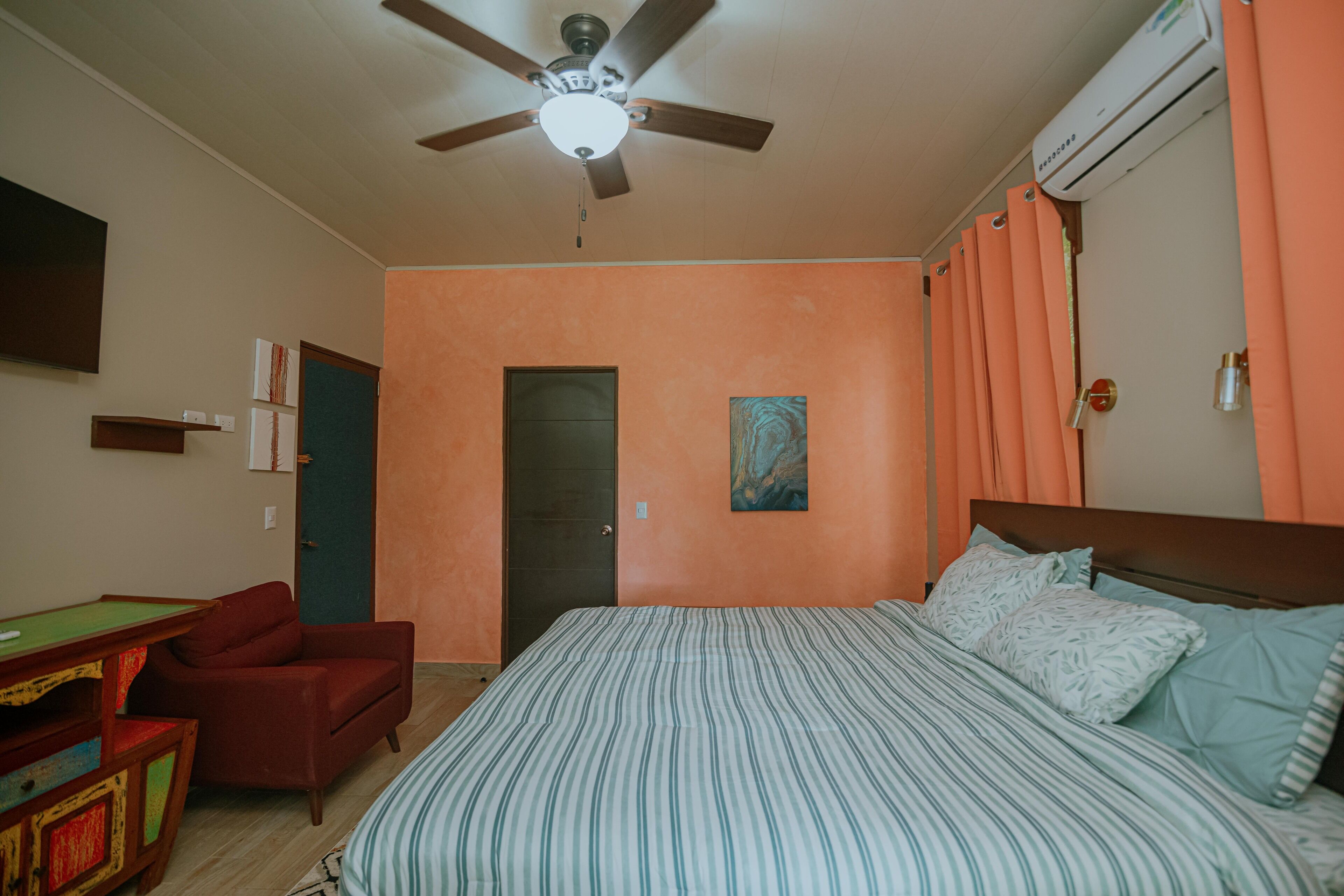 Une chambre à coucher comprenant un lit, un ventilateur de plafond, un appareil de climatisation, une porte et une petite table avec une chaise.