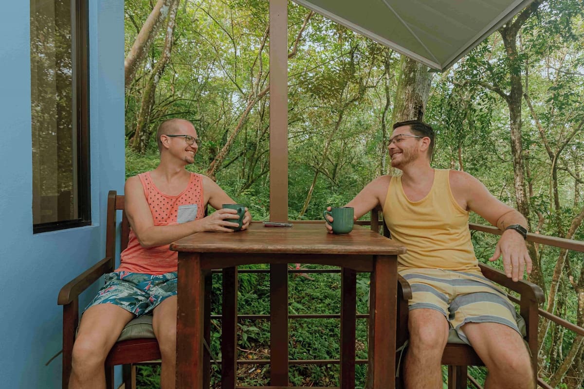 Dos hombres sentados en una mesa de madera con sillas, en una habitación con vistas a un follaje verde y denso.