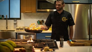 A chef in a kitchen with a stainless steel refrigerator, a gas stove with flames, and a wooden cutting board. - Oasis Diverse Adult Retreat. (Quepos)