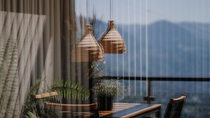 A modern interior with a wooden table, two hanging woven light fixtures, a potted plant, and a view of mountains through a large window. - Ruhiges Loft in der Natur mit Fantastischem Blick auf Meran (Dorf Tirol)