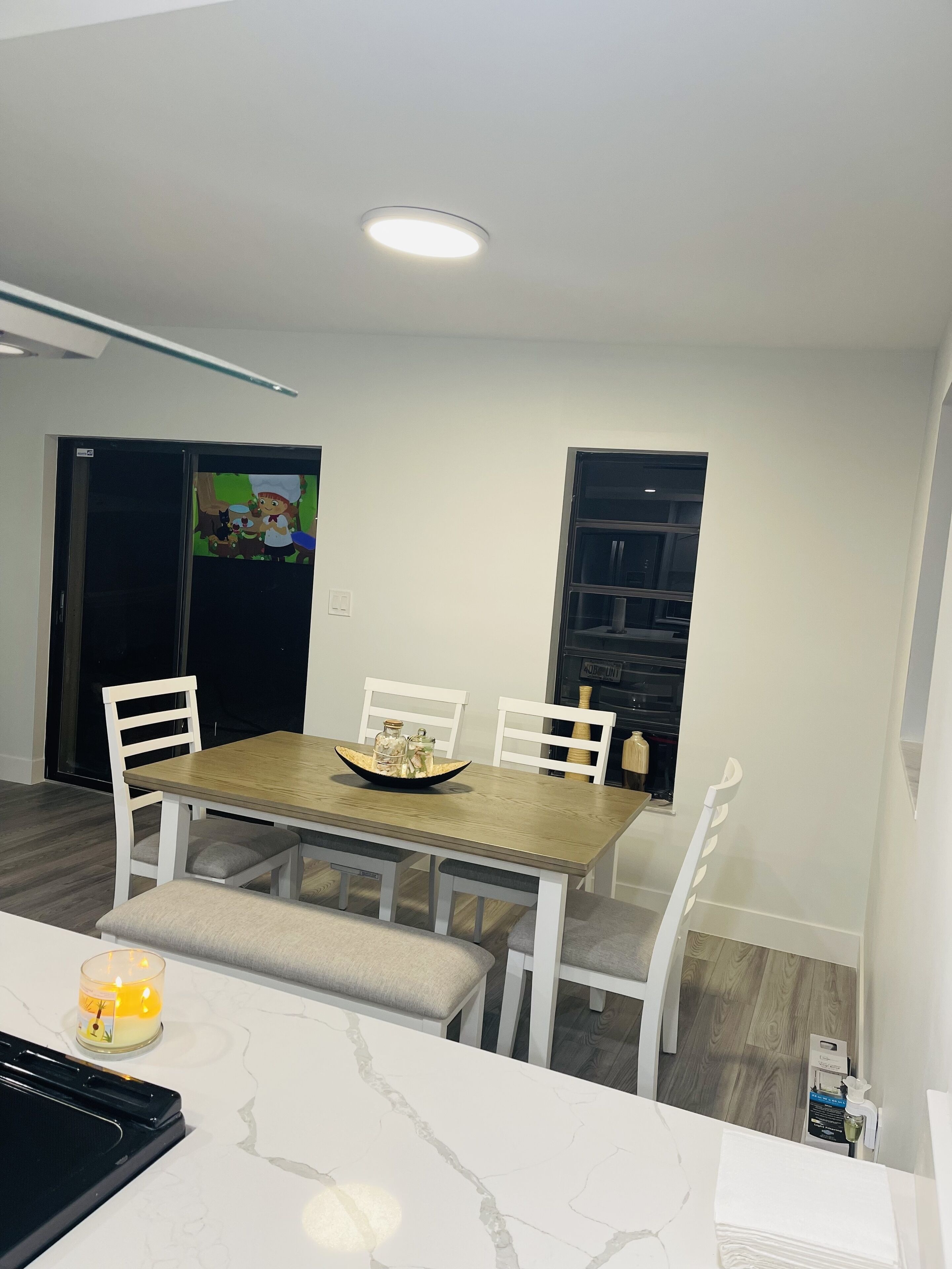 Un coin repas avec une table en bois, des chaises blanches et un banc. Une bougie est posée sur la table, ainsi qu’une petite lampe de table.