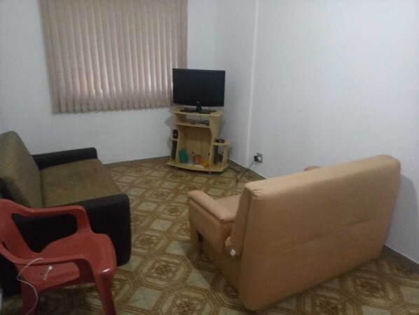 A living room with a sofa, a red plastic chair, a beige armchair, and a television on a wooden stand.