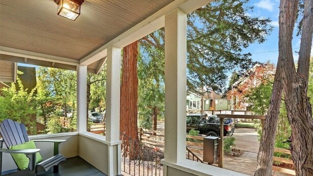 A covered porch with a table and chairs, a view of the street and houses, and a large tree in the background.