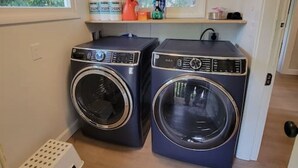 A laundry room with two front-loading washing machines, a small shelf with cleaning supplies, and a window providing natural light.