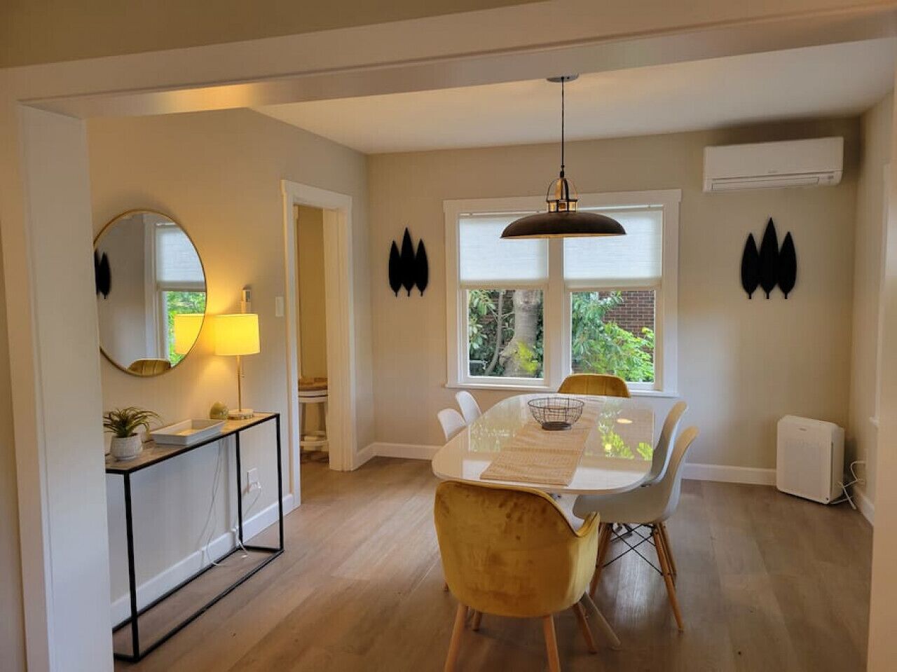 A dining area with a round table, chairs, and a pendant light. There is a window with a view of greenery outside.