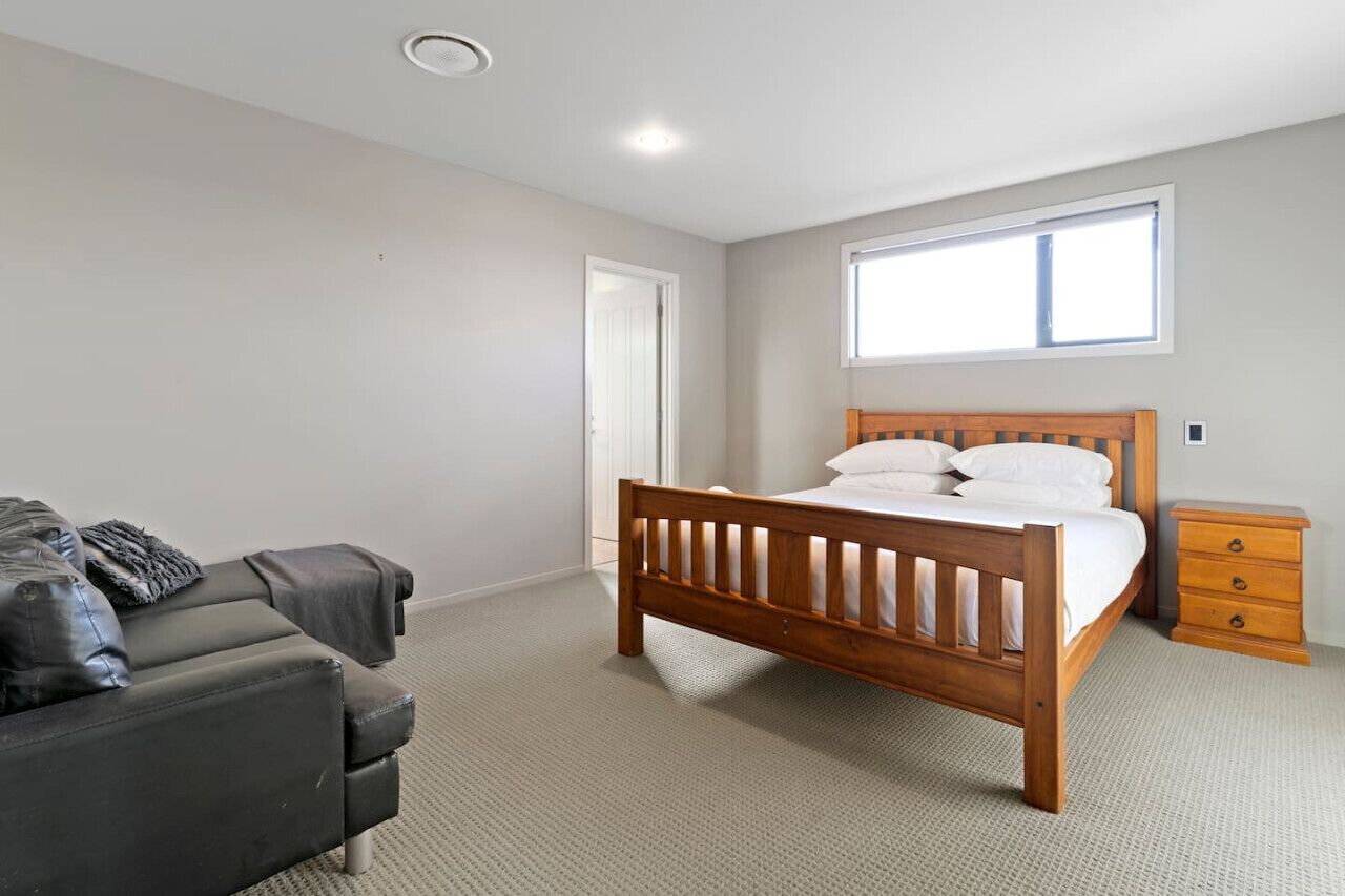 A bedroom with a wooden bed, a grey sofa, a small wooden nightstand, and a window providing natural light.