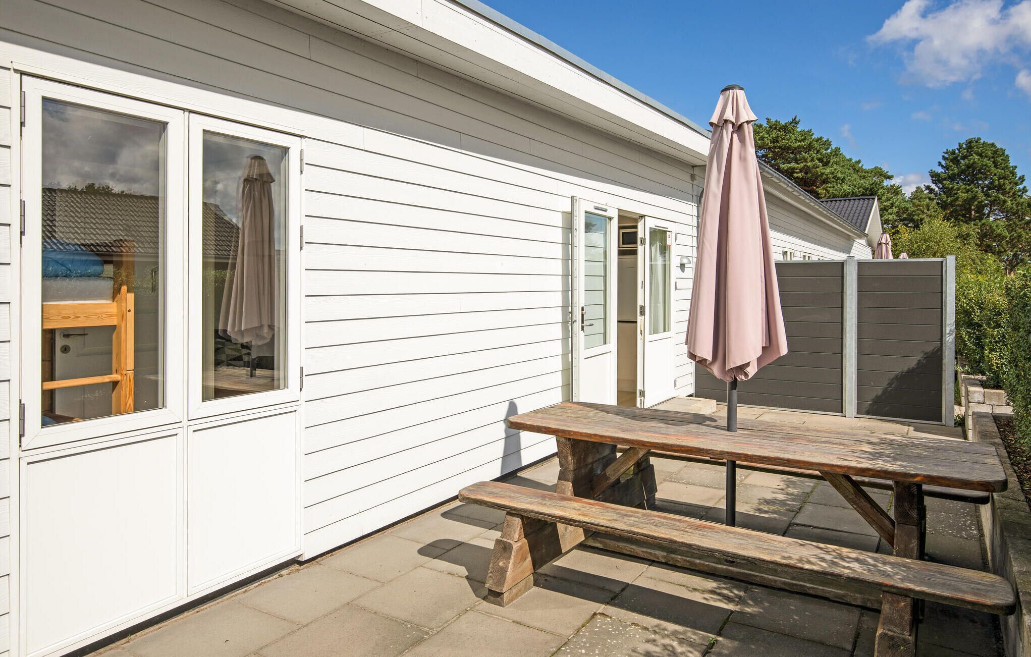 Et hvidt hus med en træterrasse, et picnicbord og en pink parasol.
