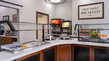 Ein Bereich mit Frühstücksbuffet, Schild mit Aufschrift 'Täglich kostenloses Frühstück', Kaffeemaschine und verschiedenen Speisen.