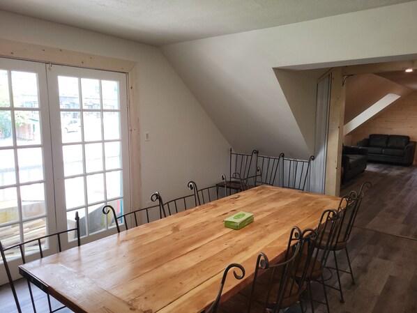 A spacious dining area with a wooden table and metal chairs, a sliding glass door leading outside, and a lofted living area with a dark-colored sofa. - The Orchard Hostel Private Bunks (Radium Hot Springs)