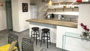 Eine moderne Küche mit einer Holzausziehtafel, zwei Barhocker, einer Pendelleuchte und einem Glastischregal mit Küchenutensilien.