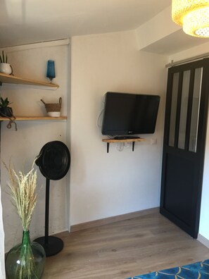 Una habitación con televisión en un soporte de pared, suelo de madera, una puerta negra y un estante pequeño con adornos.