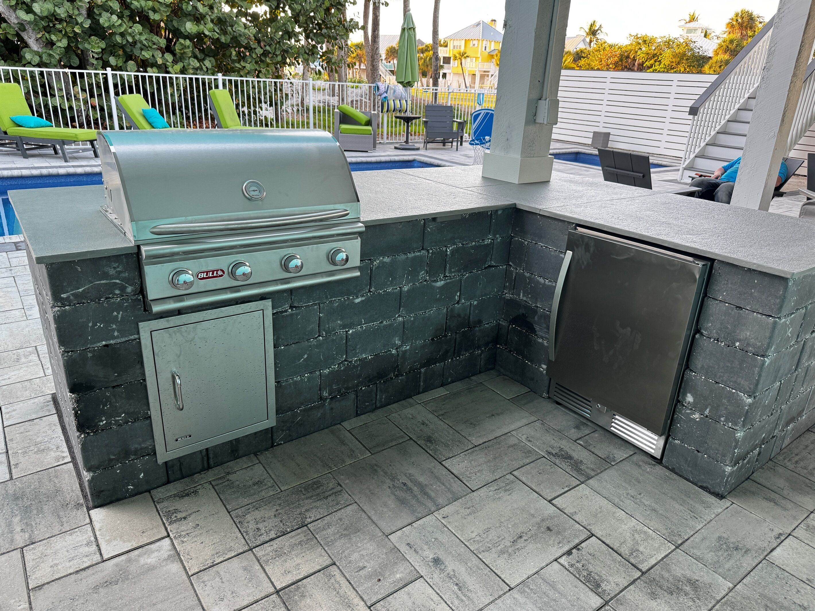 An outdoor kitchen with a grill, a sink, and a refrigerator, surrounded by a pool and lounge chairs.