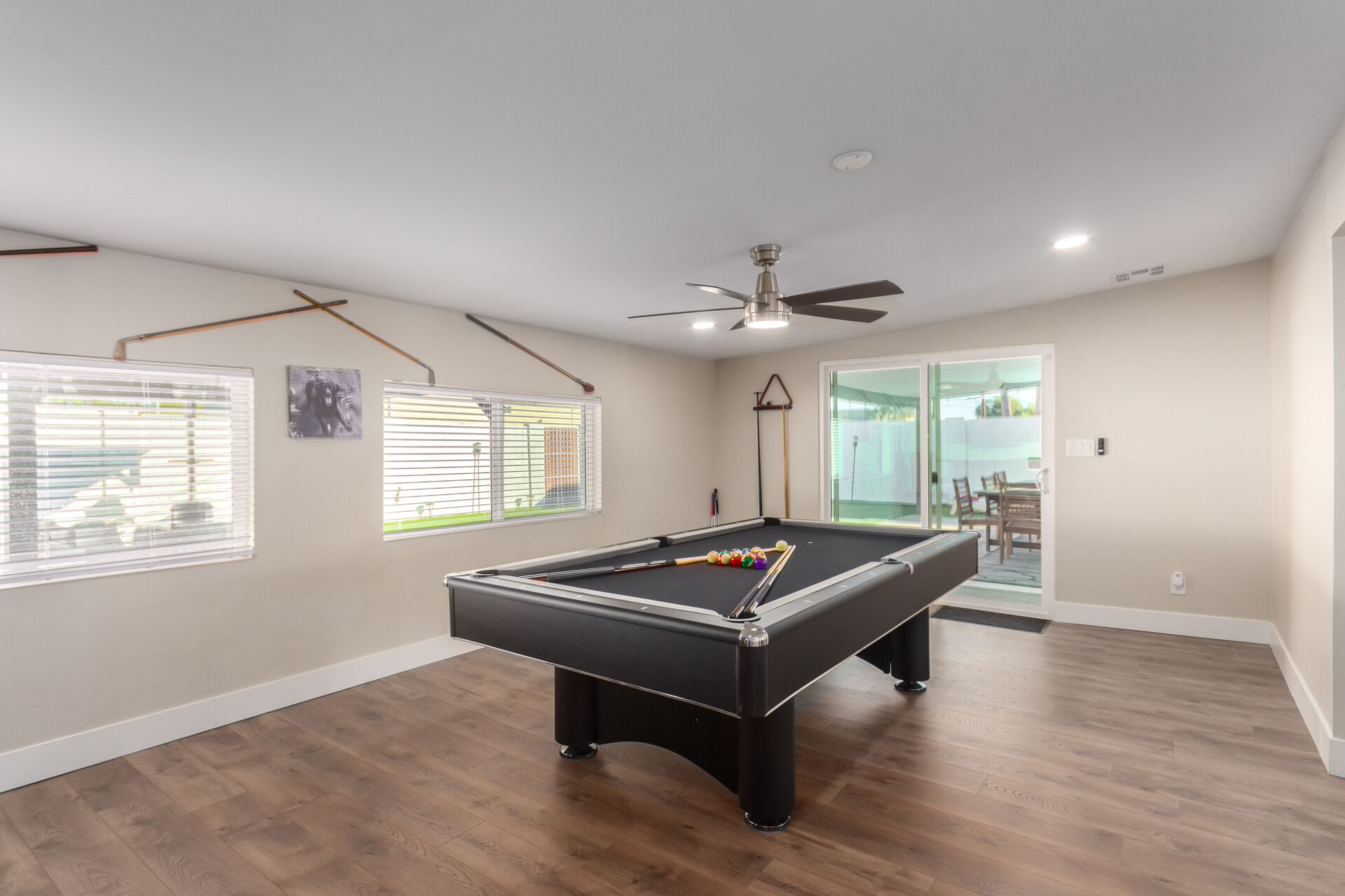 A pool table in a room with wooden flooring, a ceiling fan, and a view to an outdoor area.