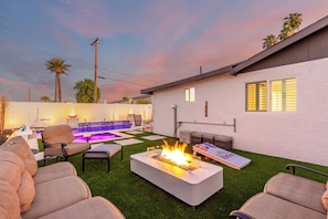 A backyard with a fire pit, outdoor seating, a pool, and a white house.