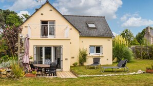 Una casa a due piani con un balcone, un patio con tavolo e sedie, e un giardino con piante e prato.
