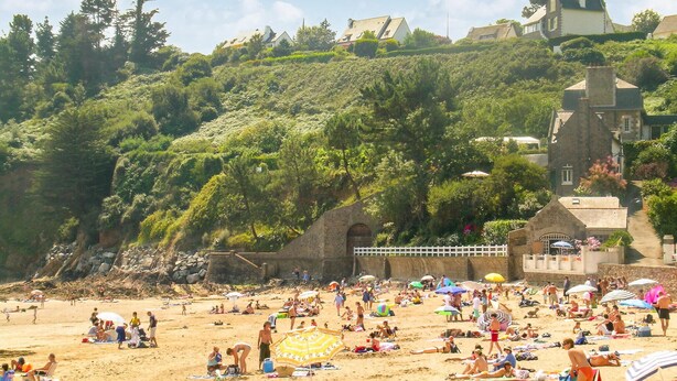 Una spiaggia con persone che prendono il sole e nuotano, circondata da rigogliosa vegetazione ed edifici storici.