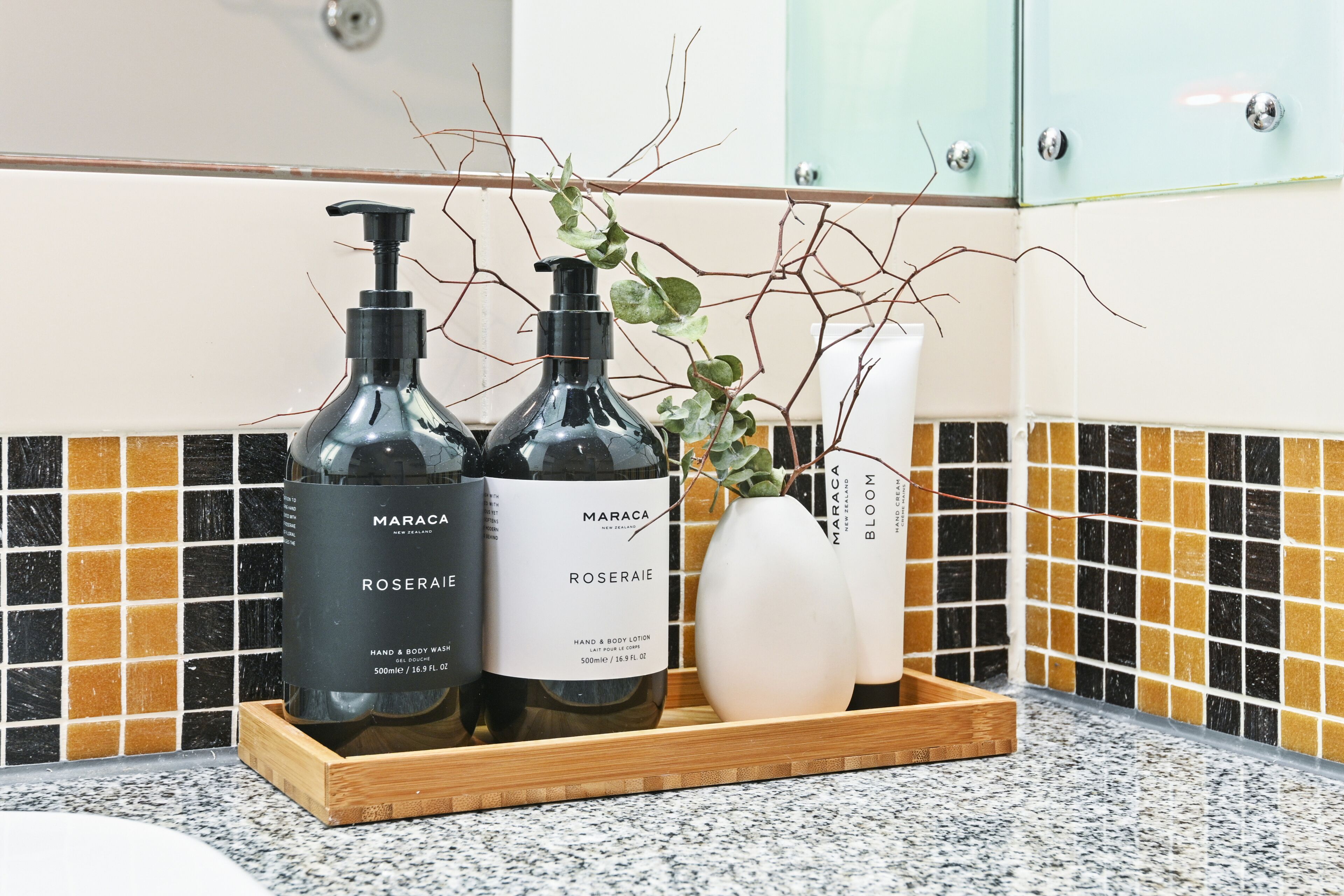 A bathroom vanity with two bottles of Maraca Roseraïe hand and body lotion, a white vase with branches, and a wooden tray.