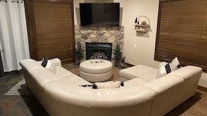 Una sala de estar con un sofá seccional color beige, un otoman redondo y un televisor de pantalla plana montado sobre una chimenea de piedra.