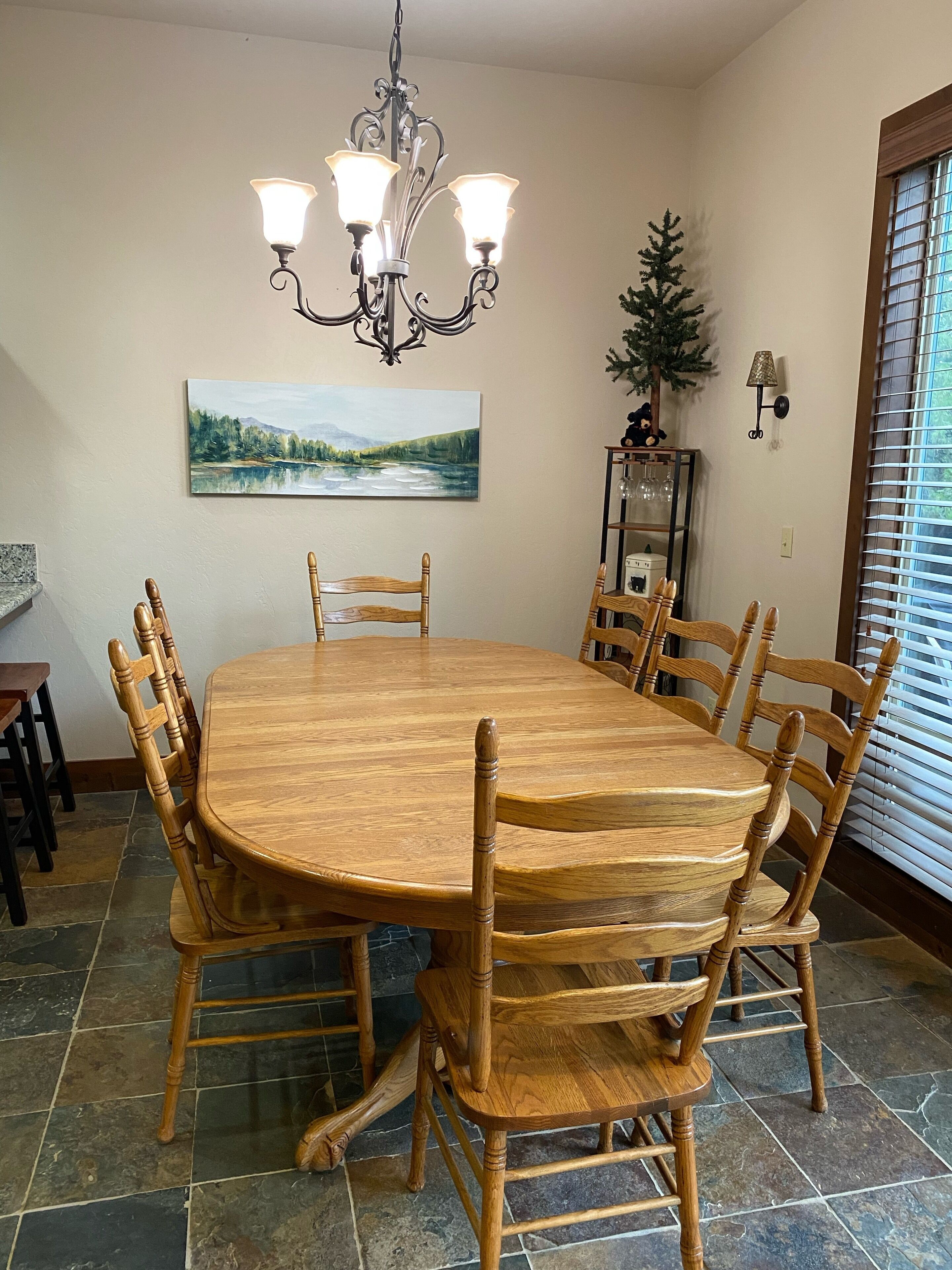 A dining room with a round wooden table and six chairs, a chandelier, a painting, a small Christmas tree, and a window with blinds.