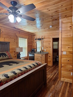 A wooden cabin interior with a bed, a ceiling fan, a kitchen area, and a bathroom.