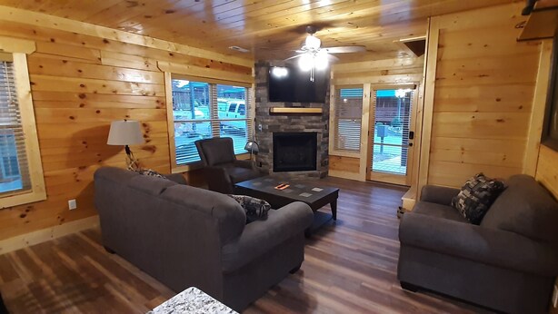 A cozy cabin-style room with wooden walls, a stone fireplace, a ceiling fan, and a large window overlooking a parking area. - Mountain Porches at Blackberry Ridge  (Near everything) (Pigeon Forge)