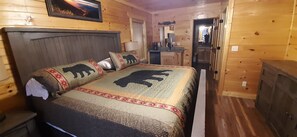 Quarto de cabana de madeira com cama, travesseiros, mesa de cabeceira, cômoda, espelho e uma pequena cozinha compacta.