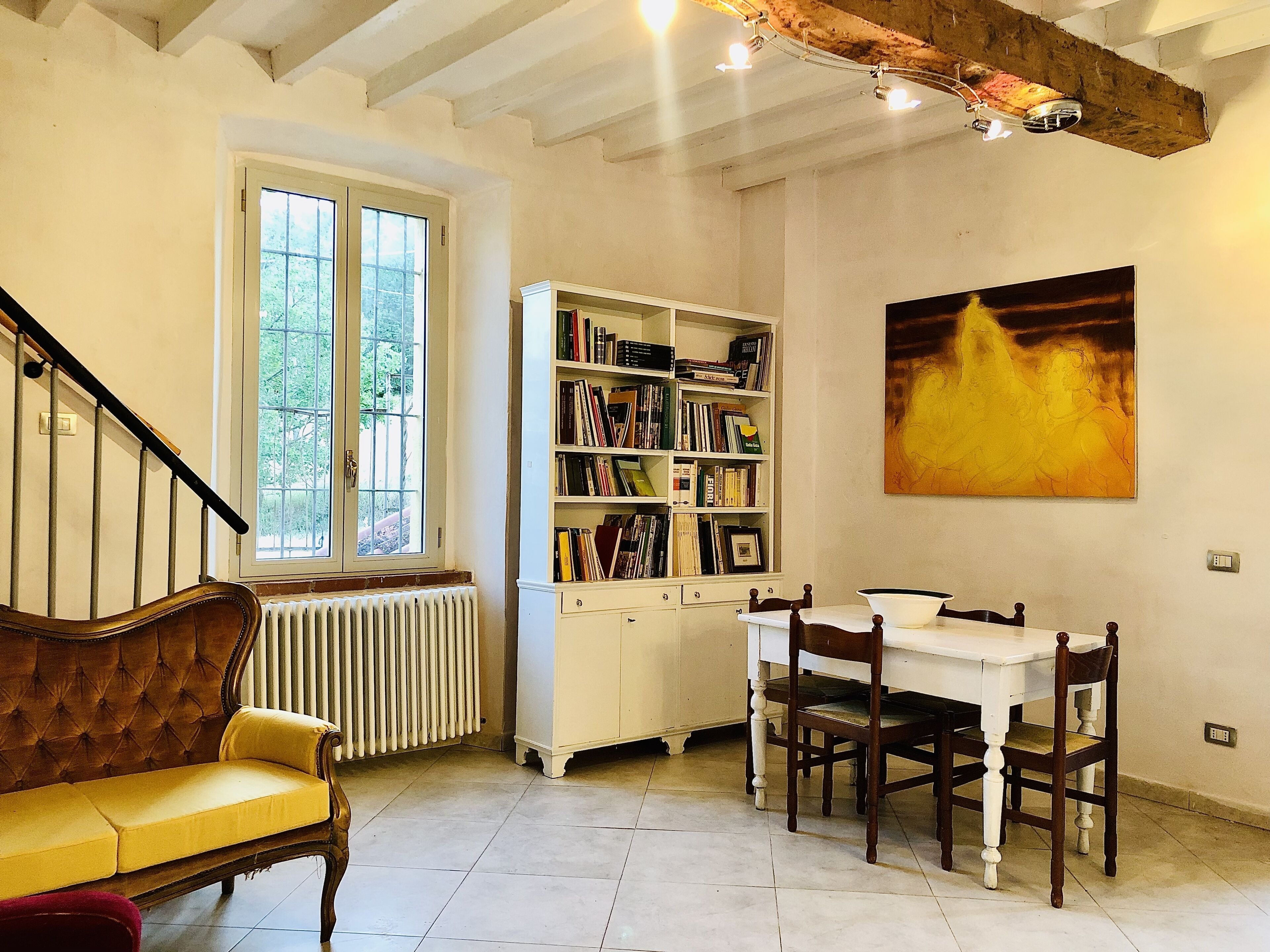 A room with a staircase, a bookshelf, a dining table with chairs, and a painting on the wall.