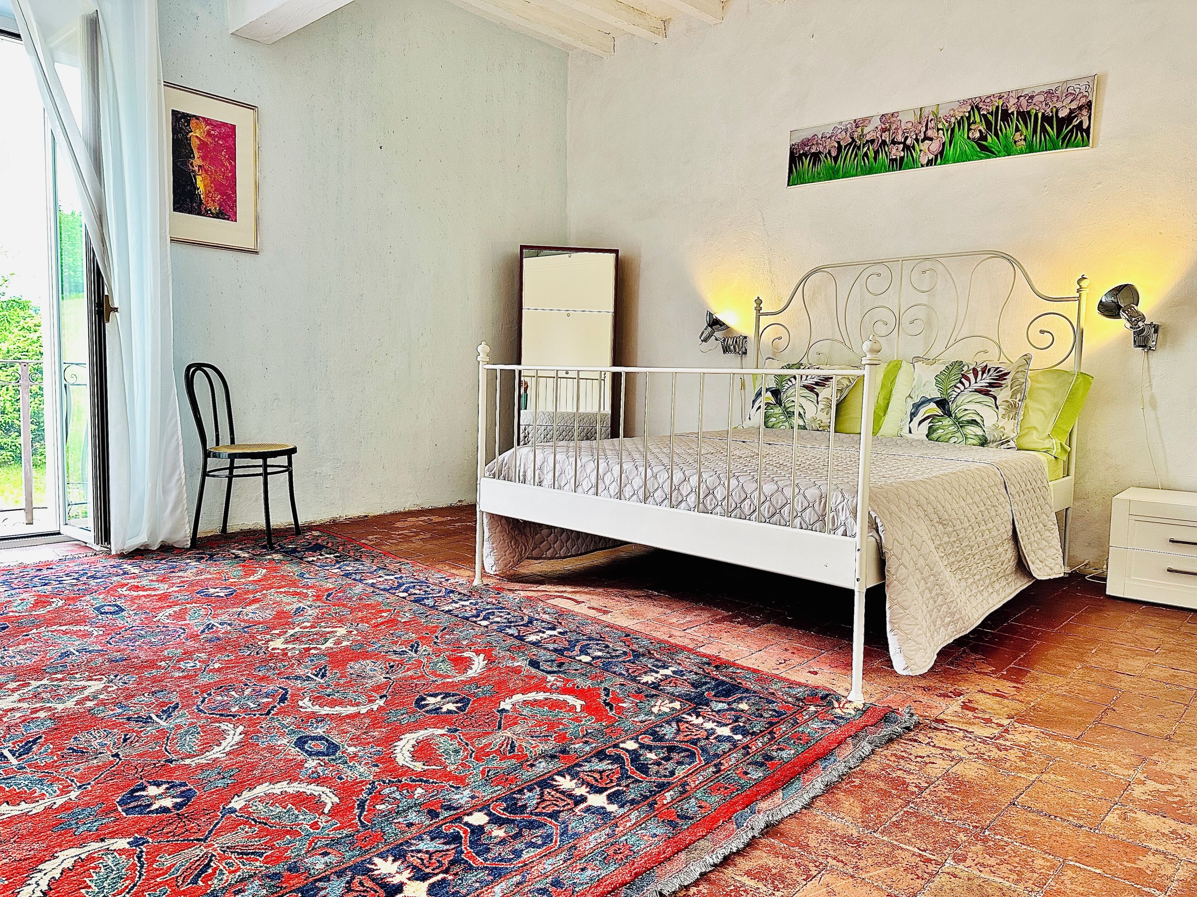 Quarto com estrutura de cama de metal branca, tapete estampado, uma cadeira e um quadro na parede.