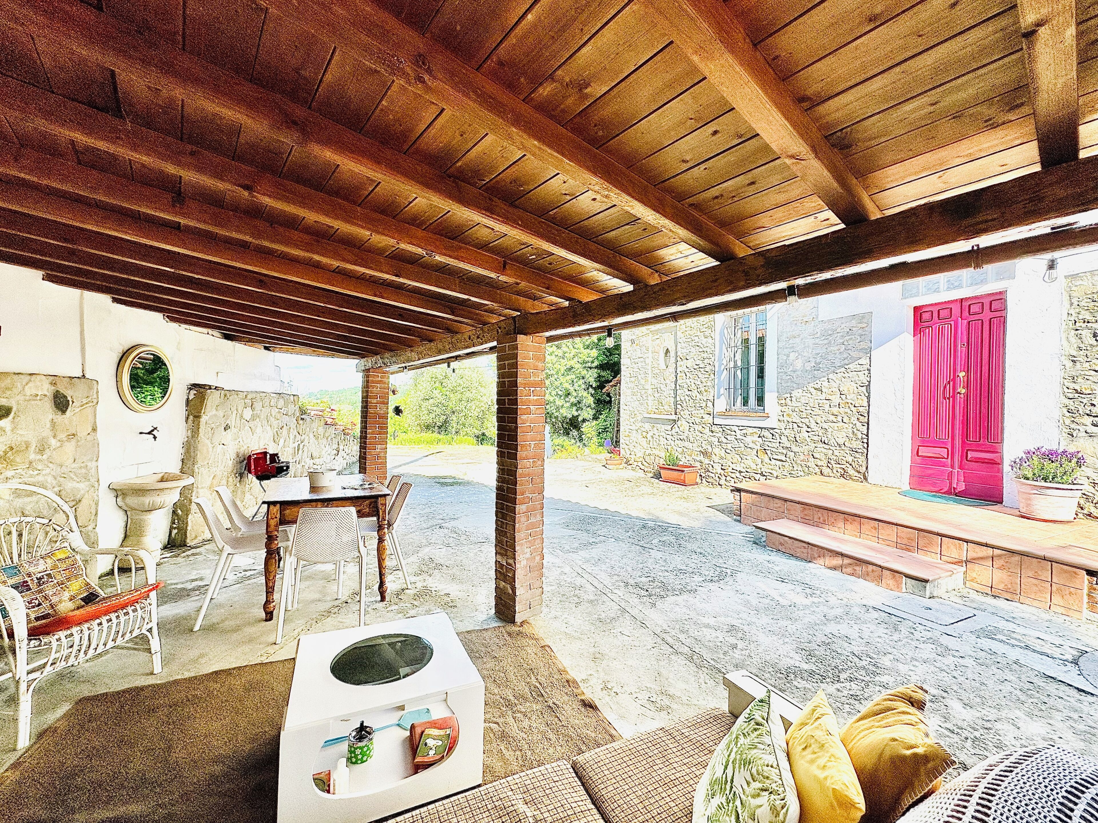 Un patio al aire libre con techo de madera, paredes de piedra, una puerta roja y una mesa blanca con sillas.