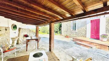 Un patio al aire libre con techo de madera, paredes de piedra, una puerta roja y una mesa blanca con sillas.