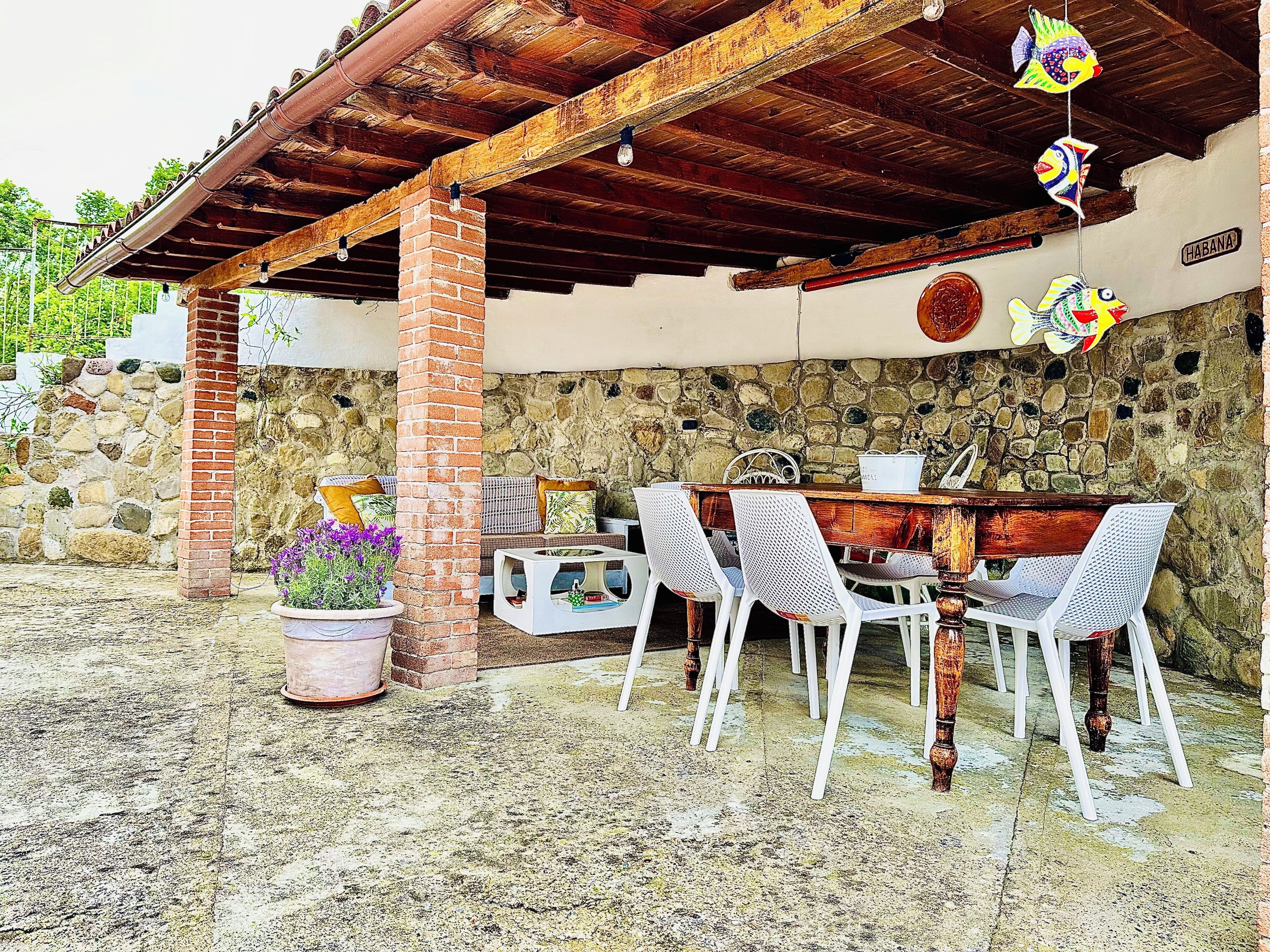 Un patio exterior con muro de piedra, pérgola de madera y una mesa con sillas.