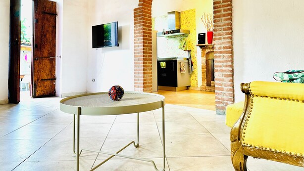 Una sala de estar con una mesa redonda, un sillón amarillo y un arco de ladrillo que da a una cocina.