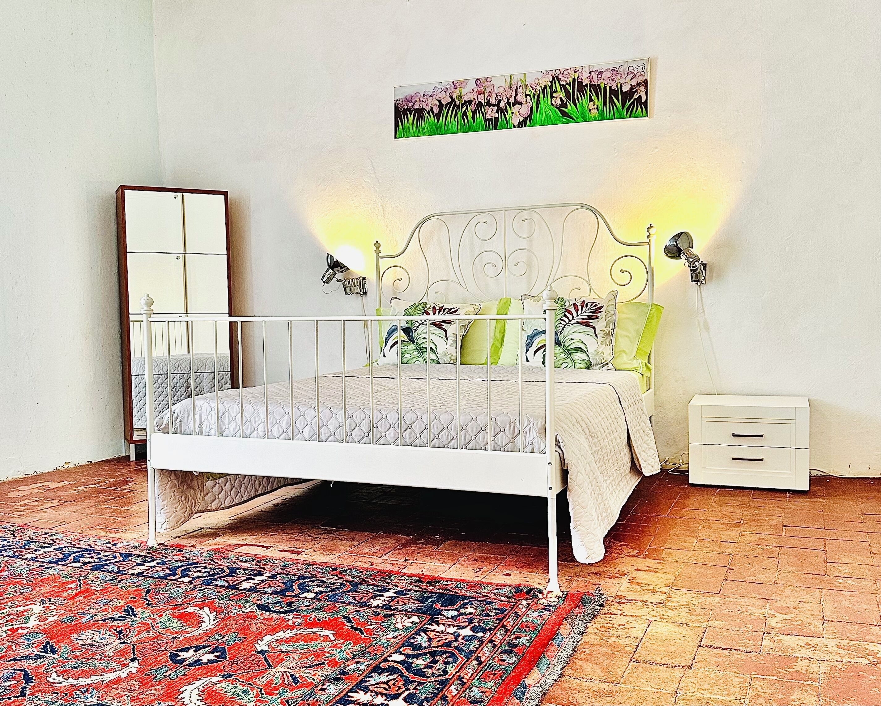 Ein Schlafzimmer mit weißem Metallbettgestell, einem gemusterten Teppich, einem Nachttisch und einer Wandlampe.