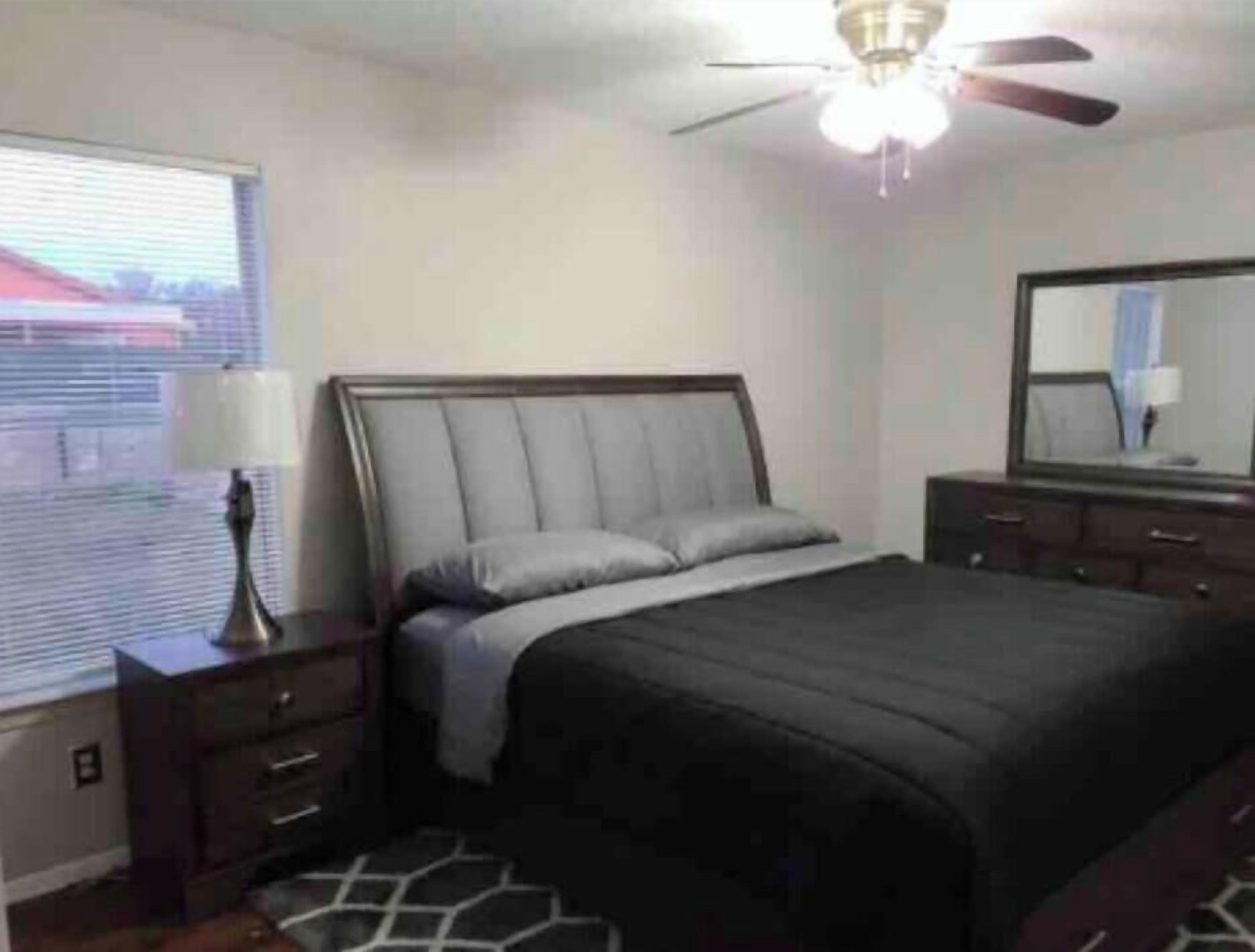 Quarto com cama, mesa de cabeceira, luminária, cômoda, espelho e ventilador de teto.