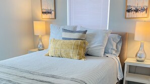 Un lit bien fait, avec une couette et des oreillers de couleur claire, une table de chevet avec une lampe et une fenêtre avec des stores.