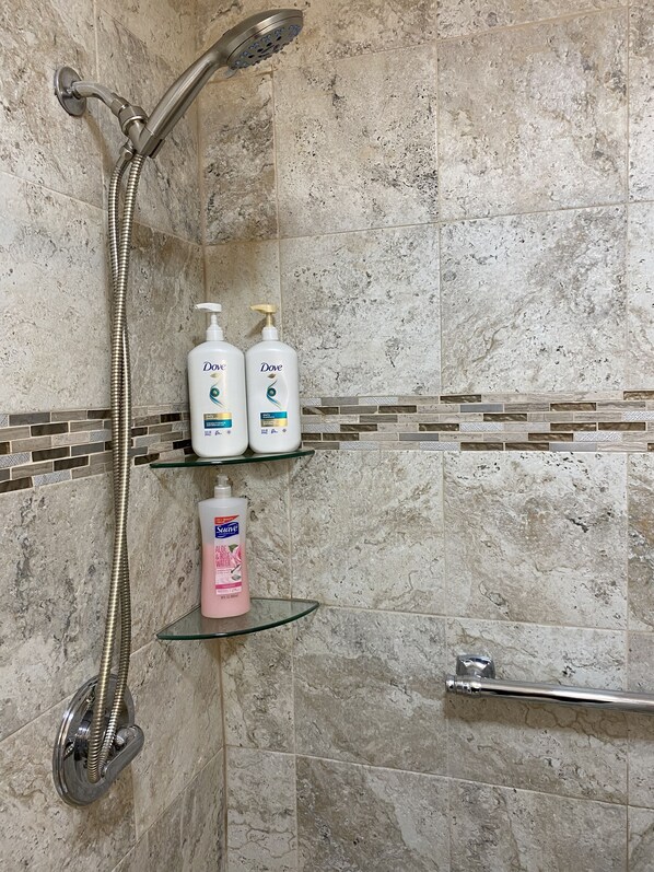 Un espace douche équipé d’un pommeau de douche à main, de deux flacons de gel douche Dove et d’un flacon d’eau de rose à l’aloe vera de la marque Suave, le tout posé sur une étagère en verre.