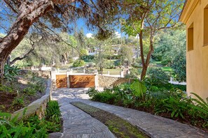 Un sendero de piedra que conduce a una puerta de madera, rodeado de abundante vegetación y un muro de piedra.