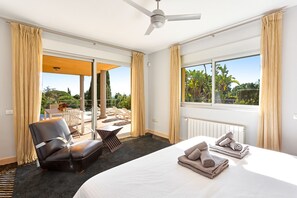 A hotel room with a bed, a chair, a table, a window with a view of greenery, and a ceiling fan. - Luxury Villa with spectaculair views Varenso Holidays (Marbella)