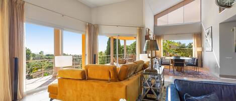 A spacious living room with a large sofa, a coffee table, and a dining area with a table and chairs. There is a balcony with a view of greenery.