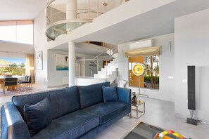 A modern living room with a blue sofa, a glass staircase, and a dining area with a view of the outdoors. - Luxury Villa with spectaculair views Varenso Holidays (Marbella)