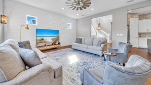 Uma sala de estar moderna com uma televisão de tela grande, um sofá branco, uma poltrona cinza e um tapete azul estampado.