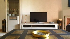 A living room with a flat-screen TV on a white entertainment unit, a round gold tray on the floor, and a glass cabinet with items on the left.
