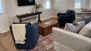 Un salon avec un fauteuil bleu foncé, un canapé de couleur claire, une table basse en bois et un téléviseur à écran plat fixé au mur.