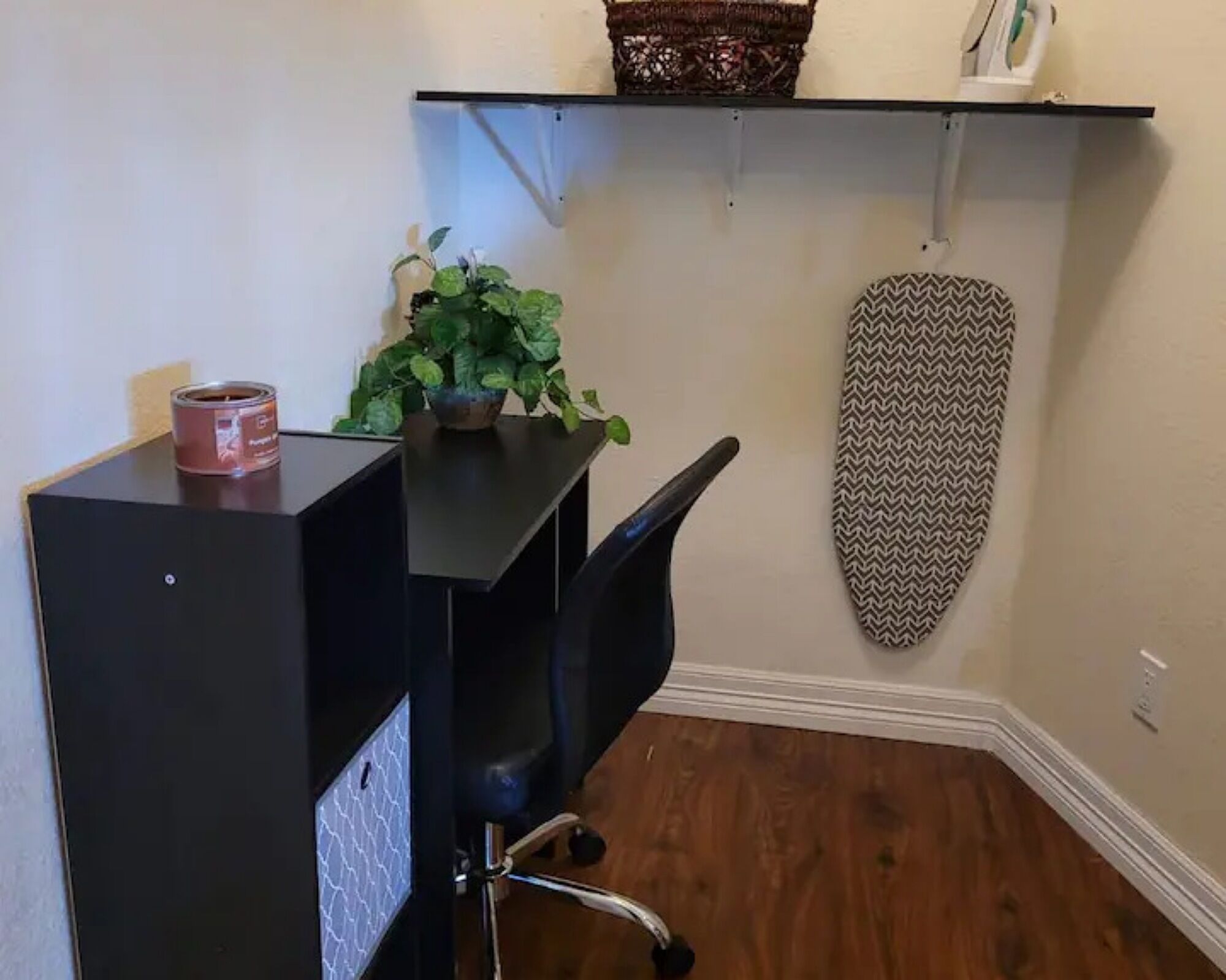 A room with a black desk, a chair, a potted plant, a shelf with a basket and a tissue box, and a patterned ironing board.