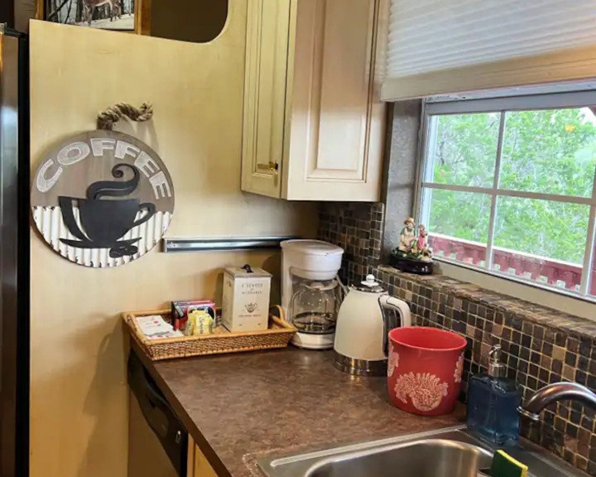 A kitchen with a coffee-themed decor, a coffee maker, a red mug, and a window with a view of greenery.