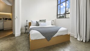 Quarto moderno com cama de madeira, roupa de cama branca, manta de padrão herringbone e vista para o horizonte urbano através de uma janela grande.