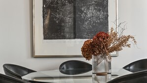 Uma sala de jantar com mesa de vidro, cadeiras pretas e um vaso com flores secas.