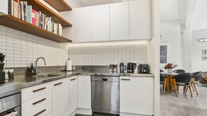 Cozinha moderna com armários brancos, máquina de lavar louça em aço inoxidável e prateleira de madeira com livros.