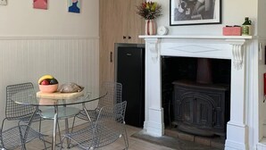 Un coin repas avec une table en verre et des chaises en fil de fer, un réfrigérateur noir, une armoire en bois et une cheminée blanche surmontée d’un tableau encadré.